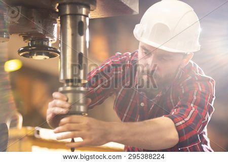 Young engineer in protective helmet bending in front of large iron drill holder while regulating working element inside it