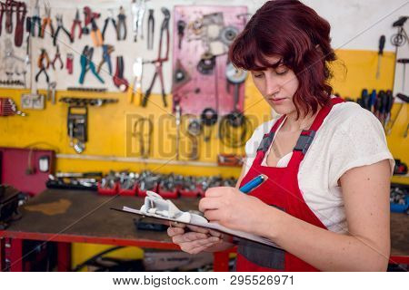 Cute, Female Mechanic In Red Work Wear Overalls Writes On Clipboard In Car Garage. Car Inspection. G