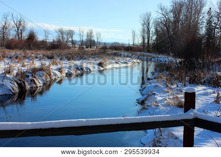 Stream Or River Running Through Snowy Landscape In Montana With Fence In Foreground