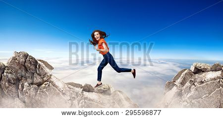 Redhead Woman Jumps Through The Gap Between Hills Over Cliff On Blue Sky Background.