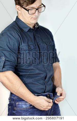 Young Handsome Caucasian Man Unzipps Blue Jeans. Man Torso, Fingerring, Blue Shirt, Glasses. Indoors