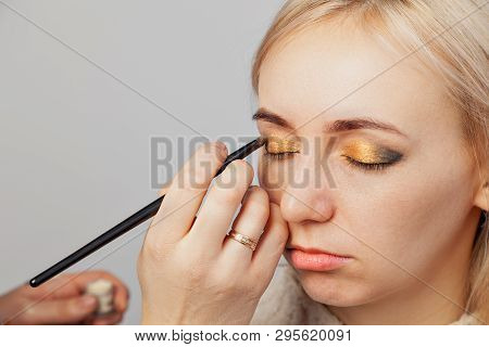 Visagiste With A Brush In One Hand Putting Makeup On The Eyes Of The Model, The Other Hand Pulls The