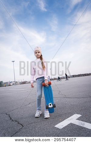 A Teenager Girl In A Pink Hat And Jeans Stands With A Pennyboard.