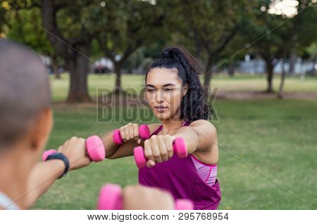 Focused woman in training session using pink dumbbells. Young determined latin woman lifting weights and working out at park. Serious girl training at outdoor gym doing hand stretching exercise.