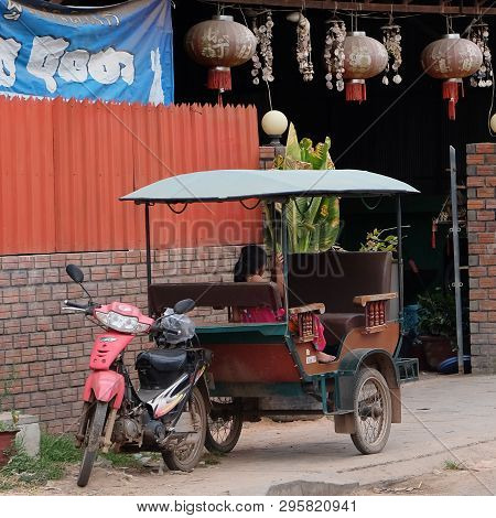 Cambodia, Siem Reap 12/08/2018 A Little Asian Girl Sits In A Moto Rickshaw Near A House With Red Lan