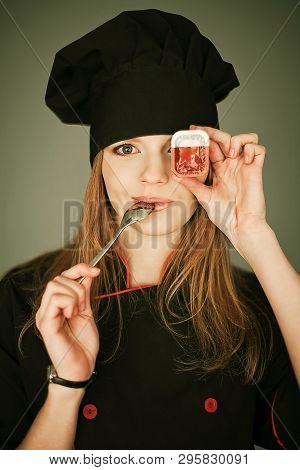 Funny Portrait Of A Joyful Lady Cook With Box Of Sauce And A Metal Spoon In The Mouth Over Gray Back