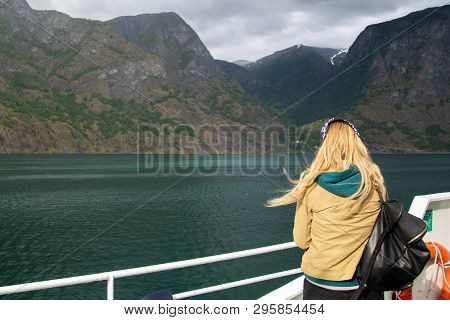 Young Woman With A Backpack On The Deck Of The Ferry Looks At The Norwegian Fjords. Woman Blonde Tou