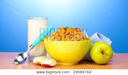 leckere Cornflakes in grüner Schale, Äpfel und Glas Milch auf Holztisch auf blauem Hintergrund