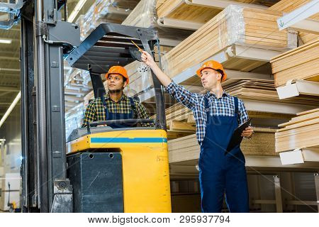 Warehouse Worker Pointing With Pencil Near Inidian Colleague Sitting In Forklift Machine