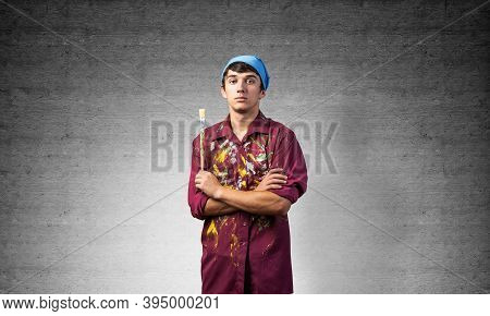 Pensive Painter Artist Standing With Folded Arms On Gray Wall Background. Portrait Of Young Man Look