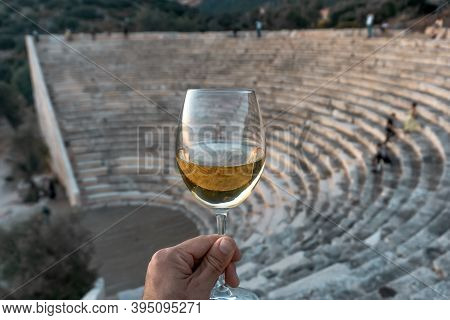 A Glass Of White Wine In A Man's Hand. Greek Mediterranean Wine Against The Backdrop Of An Antique A