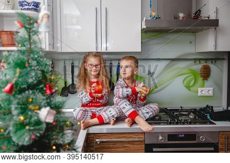 Little Laughing Twins Sit On The Table In Pajamas In The Christmas Decorated Kitchen With An Apple. 