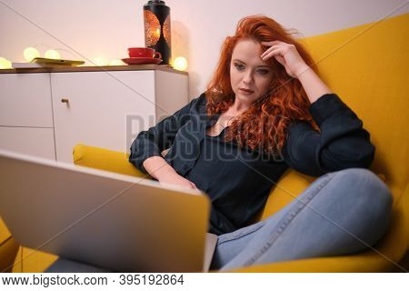 Nice beautiful lady with redhead curly hair work at the notebook sit down on the sofa at home - check on oline shops for cyber monday sales - technology woman concept for alternative office freelance