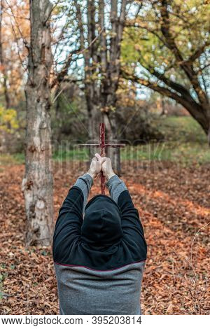 A Believing Man In A Hood Bowing His Head In Prayer And Raising The Cross Above.