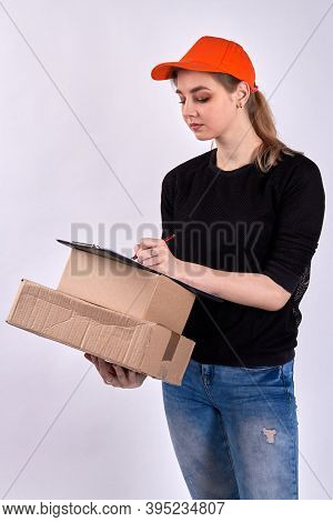 Female Courier Delivers Boxes. American Female Is Holding Pen, Clipboard With Papers Document And Pa