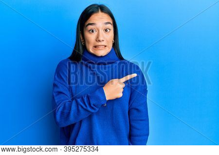 Young asian woman wearing casual winter sweater pointing aside worried and nervous with forefinger, concerned and surprised expression 