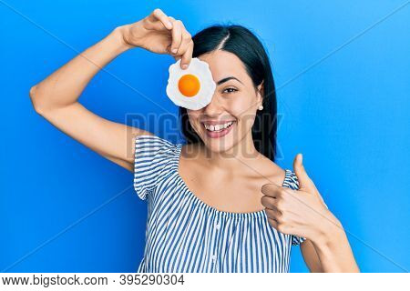 Beautiful young woman holding fried egg smiling happy and positive, thumb up doing excellent and approval sign 