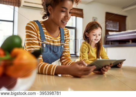 African American Woman Baby Sitter And Caucasian Cute Little Girl Reading, Using Tablet Pc, Sitting 