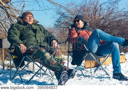 Two Young Men Rest On Camping Chairs In Campside At Sunny Day, Friends Relax In Wild, Winter Lifesty