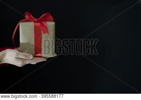 Female Hands In A Red Sweater Hold A Gift Wrapped In Craft Paper With A Wide Red Ribbon On A Black B