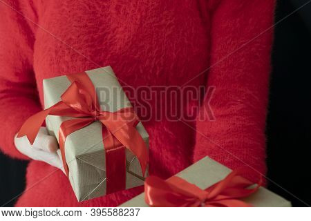 Female Hands In A Red Sweater Hold A Gift Wrapped In Craft Paper With A Wide Red Ribbon On A Black B