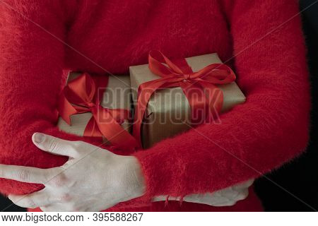 Female Hands In A Red Sweater Hold A Gift Wrapped In Craft Paper With A Wide Red Ribbon On A Black B