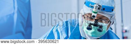 Asian Doctor Surgeon In Mask Wearing Surgical Loupes During Medical Procedure, Surgery Operating Roo