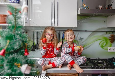 Little Laughing Twins Sit On The Table In Pajamas In The Christmas Decorated Kitchen With An Apple. 