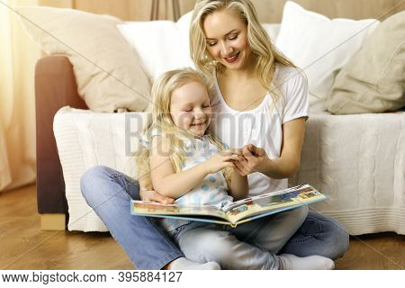 Happy Family. Blonde Young Mother Reading A Book To Her Cute Daughter While Sitting At Wooden Floor 