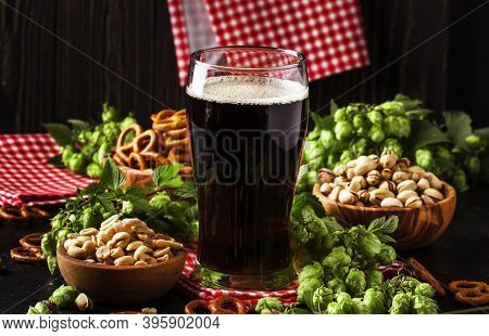 Dark German Beer Is Poured Into A Glass, Fresh Green Hops And Bowls With Salty Snacks And Nuts, Autu