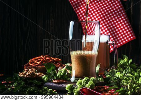 Dark German Beer Is Poured Into A Glass, Fresh Green Hops And Bowls With Salty Snacks And Nuts, Autu