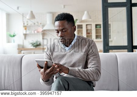 Serious African American Man Holding Digital Tablet Computer Sitting On Couch At Home. Young Adult B
