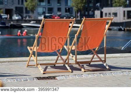 Two Orange Sun Loungers Stand On The Shore Of Blue Water, A Pond, Kayaks Float Past On A Sunny Brigh