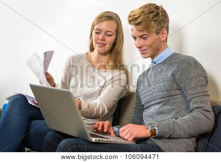 Couple, sitting on a couch, with a woman holding several bills and invoices, comparing their expenditure on the manÃ?ÃÂ¢??s laptop with their joint account cash book