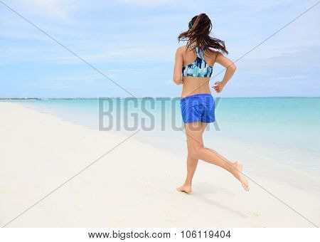 Runner training cardio running on beach. Back view of woman jogging barefoot in tropical destination barefoot in activewear blue sports bra and active shorts living a healthy life.
