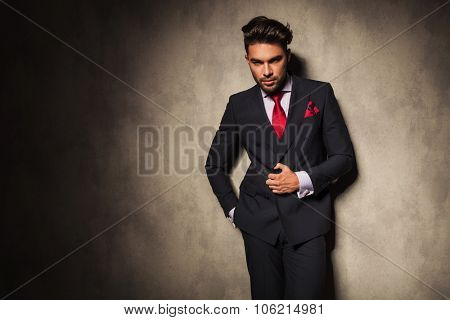 Attractive young business man closing his jacket while leaning on a grey wall.