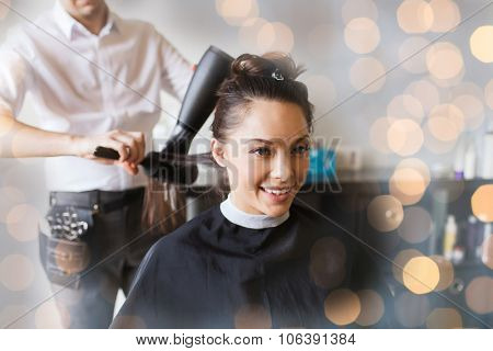 beauty, hairstyle and people concept - happy young woman and hairdresser with fan making hot styling at hair salon over holidays lights