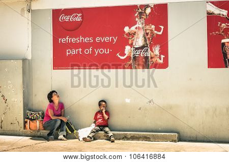 Namibian Mother And Son Sitting On Floor Under A Coca Cola Billboard