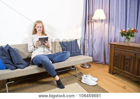 Young woman, sitting comfortably on a modern sofa, surfing on a portable device in a living room, with the curtains drawn