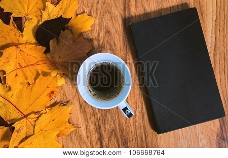 Old black book and cup of coffee on the desk among yellow leafs