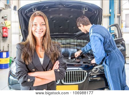 Young business woman, smiling and looking into the camera, whilst a mechanic tends to the maintenance of her car in the background at a dedicated professional garage