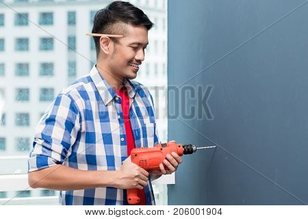 Asian do-it-yourself handyman drilling at wall in skyscraper building
