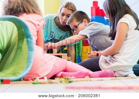 Cute pre-school boy cooperating with his colleagues at the construction of a structure, made of wooden toy blocks under the guidance of a young kindergarten teacher