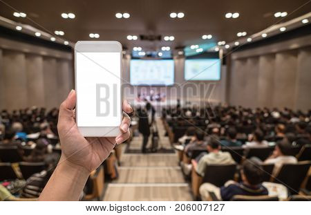 Female hand holding mobile phone when Live via social network over Abstract blurred photo of conference hall or seminar room with attendee background business technology concept