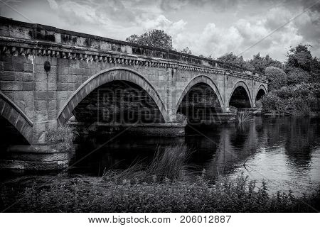 Stone Bridge over The River Trent between Repton and Willington Derbyshire. Showing the river cloudy sunny blue sky green folage foreground reflection of bridge in water.