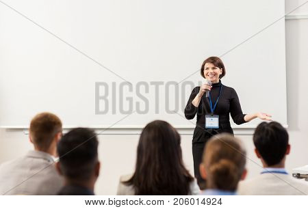 business, education and people concept - smiling businesswoman or teacher with microphone talking to group of students at conference presentation or lecture