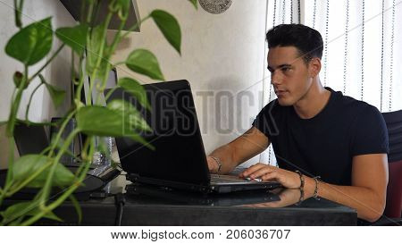 Attractive Young Man with Serious Expression, with Laptop and Desktop Comptuers, Very Busy Working on his Start-up Business - Young Male College or University Student Doing Homework on Sofa