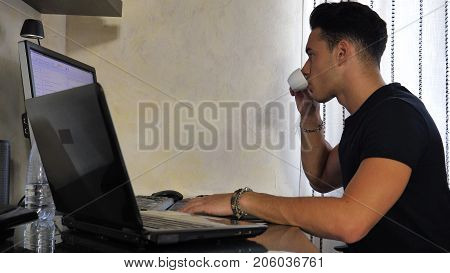 Attractive Young Man with Serious Expression, with Laptop and Desktop Comptuers, Very Busy Working on his Start-up Business Drinking Espresso Coffee - Young Male College or University Student Doing Homework on Sofa