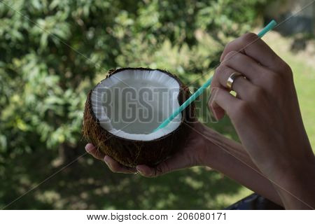 Closeup woman's hand holding coconut with green starw isolated over blurred green tropical garden background