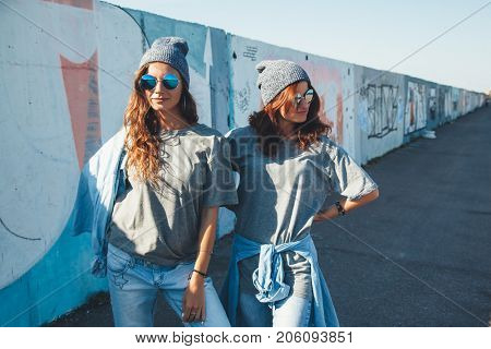 Two models wearing plain gray t-shirts and hipster sunglasses posing against street wall. Teen urban clothing style, same look. Mockup for tshirt print store.